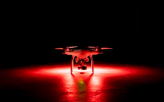 A drone is positioned on the floor, illuminated by a striking red light in an otherwise dark environment.