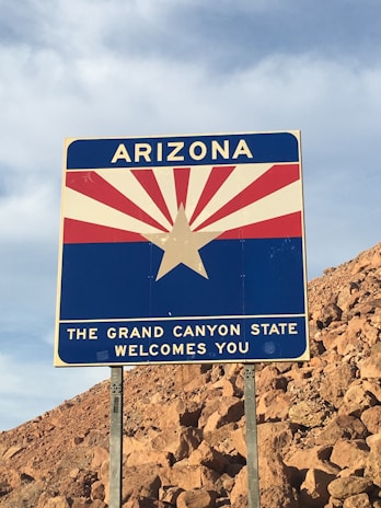 A road sign welcomes travelers to Arizona, featuring a graphic of a rising sun with red and white rays and a star in the center. The sign includes bold uppercase text, highlighting Arizona as the Grand Canyon State.