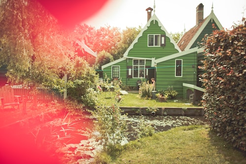A charming green wooden house with white trim sits amidst a lush garden. The scene includes various plants and a small pond bordered by stones. The architecture features a pointed roof with decorative elements. Sunlight filters through, casting warm tones. A person is visible near the entrance, adding a sense of human presence. The foreground showcases a bright red lens flare, adding a unique hue to the image.