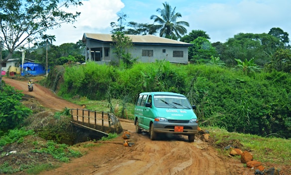 A muddy rural road features a small green bus labeled as a school bus, heading over a narrow bridge. Surrounding the road are lush green plants and trees, including a tall palm tree near a modest house built from concrete blocks. In the distance, a motorbike is seen on a dirt path. The area appears to be a tropical setting with vibrant vegetation.