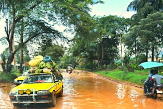 A yellow car is loaded with various goods, including bunches of bananas, and navigating a muddy, wet road surrounded by lush green trees. In the background, a motorbike with two people rides through the same road while holding an umbrella.