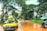 A yellow car is loaded with various goods, including bunches of bananas, and navigating a muddy, wet road surrounded by lush green trees. In the background, a motorbike with two people rides through the same road while holding an umbrella.