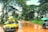 A yellow car is loaded with various goods, including bunches of bananas, and navigating a muddy, wet road surrounded by lush green trees. In the background, a motorbike with two people rides through the same road while holding an umbrella.