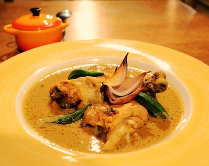 A dish featuring pieces of roasted chicken served in a creamy gravy, garnished with sliced onions and green vegetables. The meal is presented on a yellow-bordered plate, with a small orange pot in the background sitting on a wooden table.