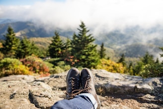 A pair of hiking boots is visible in the foreground, resting on a rocky surface. In the background, a scenic view of a lush, green forested landscape is partially shrouded in mist. The sky above is bright with clouds scattering the sunlight.