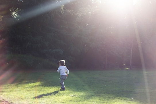 A child is running on a grassy field with sunlight streaming through the trees in the background, creating a serene and peaceful atmosphere. The light creates lens flare, adding a dreamy effect to the image.