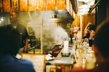 A cozy, bustling Japanese izakaya with a warm atmosphere. Patrons sit at a counter enjoying food and drinks, with a chef standing behind the counter. There is visible smoke rising from a grill, adding to the lively environment. Traditional Japanese decorations, such as hanging lanterns with Kanji characters, adorn the space.