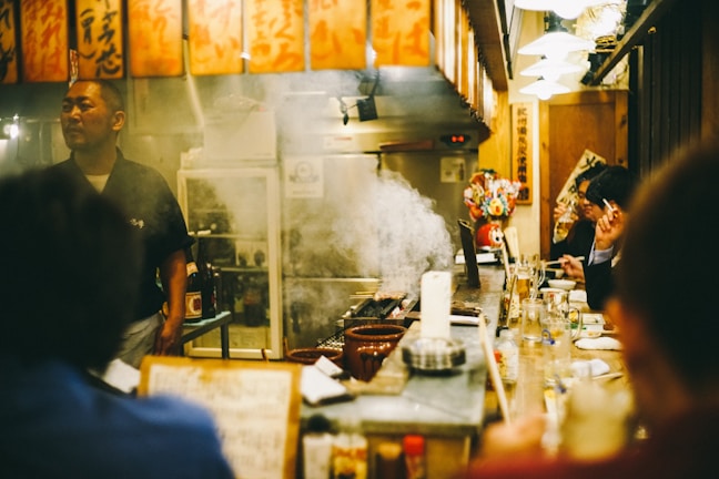 A cozy, bustling Japanese izakaya with a warm atmosphere. Patrons sit at a counter enjoying food and drinks, with a chef standing behind the counter. There is visible smoke rising from a grill, adding to the lively environment. Traditional Japanese decorations, such as hanging lanterns with Kanji characters, adorn the space.
