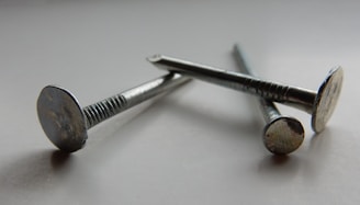 Three metallic nails are arranged diagonally on a smooth, possibly reflective surface. The nails have a shiny, silver appearance, showing details of their flat heads and textured shafts. The lighting creates soft shadows and highlights, enhancing the metallic texture.
