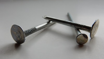 Three metallic nails are arranged diagonally on a smooth, possibly reflective surface. The nails have a shiny, silver appearance, showing details of their flat heads and textured shafts. The lighting creates soft shadows and highlights, enhancing the metallic texture.