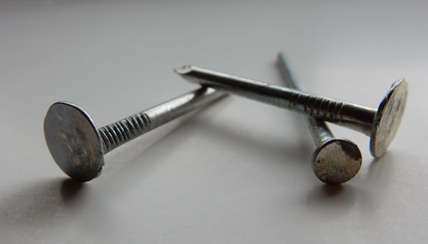 Three metallic nails are arranged diagonally on a smooth, possibly reflective surface. The nails have a shiny, silver appearance, showing details of their flat heads and textured shafts. The lighting creates soft shadows and highlights, enhancing the metallic texture.