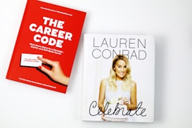 Two books are placed on a white surface. The book on the left is titled 'The Career Code' with a red cover, featuring a hand holding a business card, authored by Hillary Kerr and Katherine Power. The book on the right is titled 'Celebrate' with a white cover, displaying a photograph of a woman holding a cake.