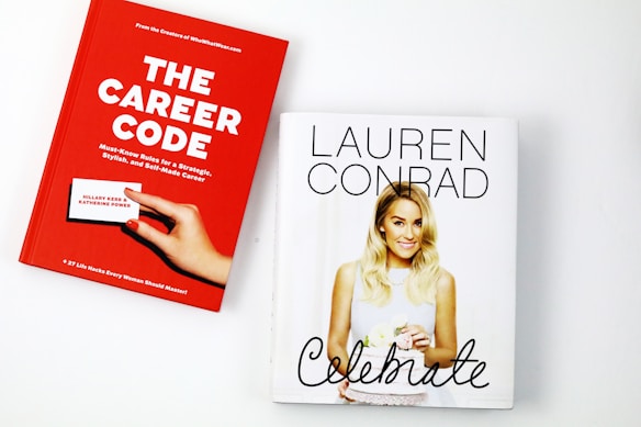 Two books are placed on a white surface. The book on the left is titled 'The Career Code' with a red cover, featuring a hand holding a business card, authored by Hillary Kerr and Katherine Power. The book on the right is titled 'Celebrate' with a white cover, displaying a photograph of a woman holding a cake.