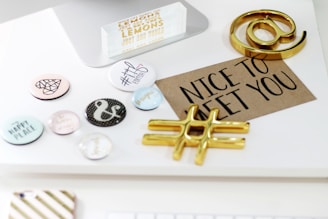 A desktop arrangement with decorative items including a series of motivational and hashtag-themed buttons, a brown card with 'NICE TO MEET YOU' printed on it, and metallic symbolic objects resembling an at symbol and a hashtag. The setting has a clean and modern aesthetic.
