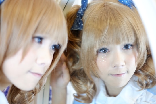 A person with light blonde hair styled in curls and adorned with small stars on their cheek is looking into a mirror. The reflection also shows their face with large, expressive eyes, giving a gentle and contemplative look. The hair is tied with a blue bow, and they are wearing a white garment.