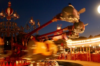 A brightly lit amusement park ride with flying elephant-shaped gondolas against a darkening evening sky. The ride is in motion, creating a sense of excitement and movement. Crowds of people are waiting in line, surrounded by ornate and colorful decorations.