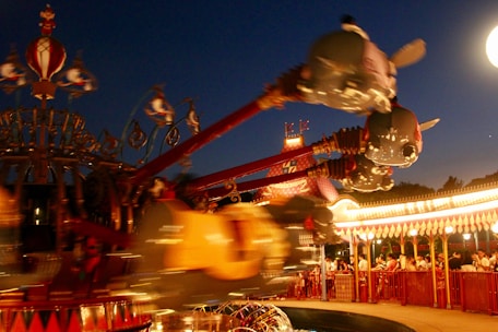 A brightly lit amusement park ride with flying elephant-shaped gondolas against a darkening evening sky. The ride is in motion, creating a sense of excitement and movement. Crowds of people are waiting in line, surrounded by ornate and colorful decorations.