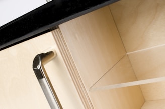 A close-up view of a wooden cabinet with a metallic handle. The cabinet door is partially open, revealing an empty shelf inside. The countertop above is dark, contrasting with the light wood of the cabinet.