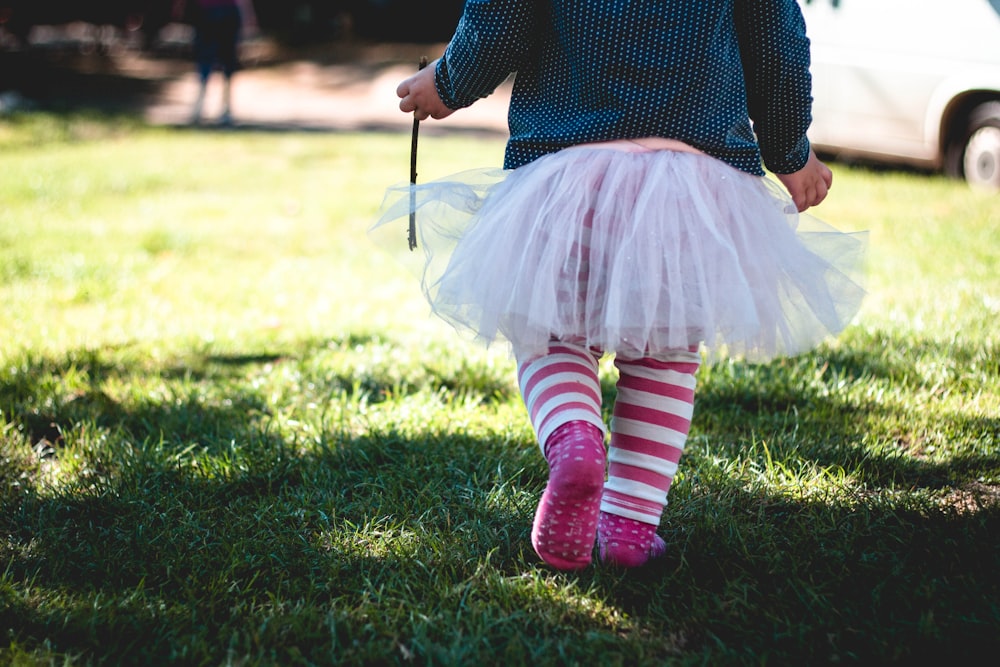青緑と白の水玉模様の長袖シャツと白いチュチュのスカートを着た幼児の女の子が昼間、緑の芝生の上を歩いています