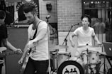 A black and white image depicting a small band performing in an outdoor setting. The scene shows a guitarist in the foreground tuning his instrument and a drummer in the background actively playing. They appear focused on their music, with musical equipment surrounding them.
