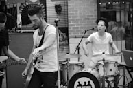 A black and white image depicting a small band performing in an outdoor setting. The scene shows a guitarist in the foreground tuning his instrument and a drummer in the background actively playing. They appear focused on their music, with musical equipment surrounding them.