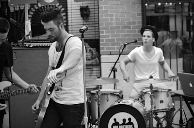 A black and white image depicting a small band performing in an outdoor setting. The scene shows a guitarist in the foreground tuning his instrument and a drummer in the background actively playing. They appear focused on their music, with musical equipment surrounding them.