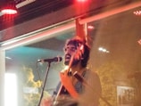 A musician passionately performing in a dimly lit venue, playing an acoustic guitar and singing into a microphone. The setting includes visible reflections on a window and an overall warm glow from red lighting.