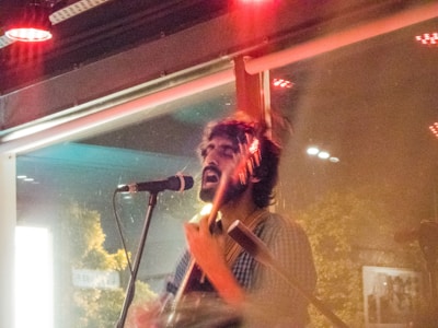 A musician passionately performing in a dimly lit venue, playing an acoustic guitar and singing into a microphone. The setting includes visible reflections on a window and an overall warm glow from red lighting.
