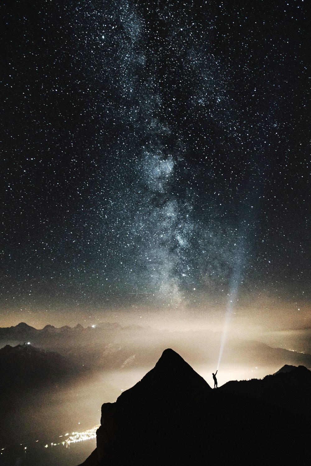 silhouette of person on top of mountain pointing flashlight on sky filled with stars at night time