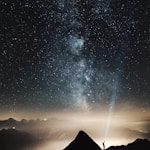 silhouette of person on top of mountain pointing flashlight on sky filled with stars at night time