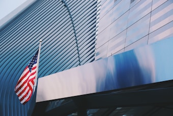 A tall flagpole with the American flag is positioned in front of a modern, architecturally complex building with a facade characterized by sleek, horizontal metal lines and reflective glass panels. The structure's design is both futuristic and artistic, showcasing a blend of straight and curved lines.