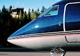 The nose of a sleek, modern airplane with a dark blue and gray color scheme is prominently visible. The background features a control tower surrounded by green trees under a clear blue sky.