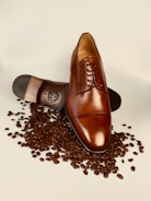 A polished brown leather dress shoe lies atop scattered coffee beans, with its sole partially visible displaying intricate detailing.