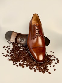 A polished brown leather dress shoe lies atop scattered coffee beans, with its sole partially visible displaying intricate detailing.