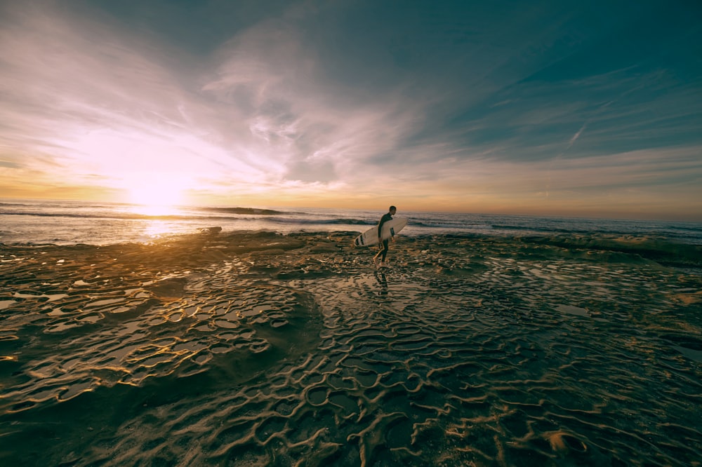 man carrying white surfboard while walking on shoreline at daytime