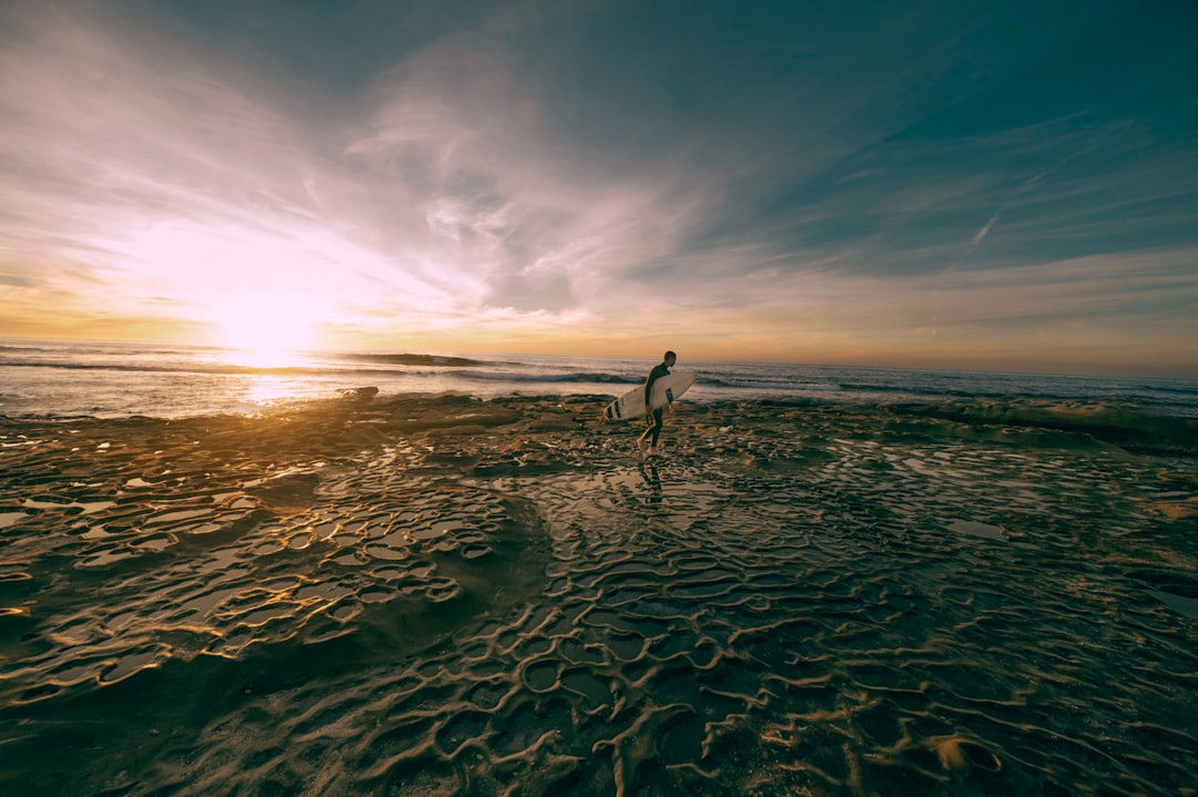 man carrying white surfboard while walking on shoreline at daytime