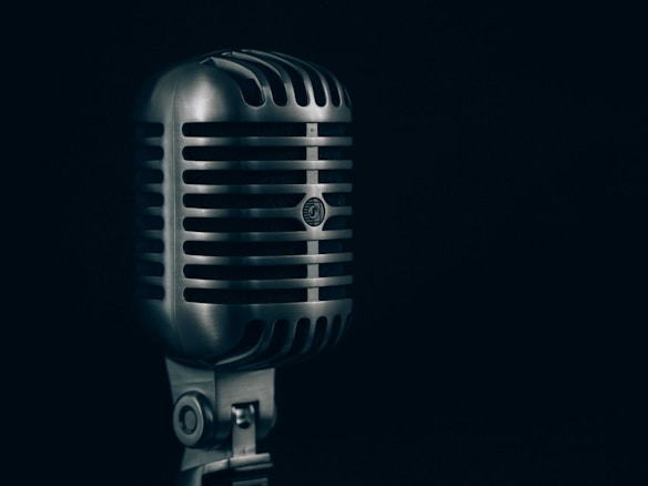 A vintage-style silver microphone stands prominently against a dark background. The microphone features a classic grille design with horizontal lines and a robust construction, evoking a sense of nostalgia and timelessness.