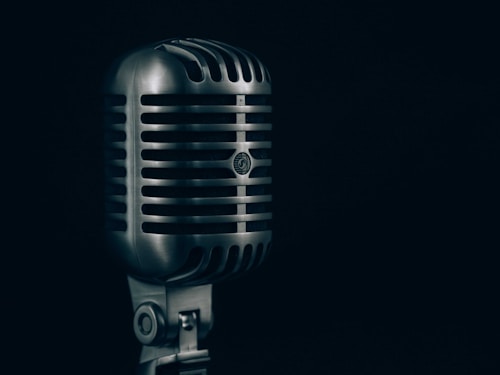 A vintage-style silver microphone stands prominently against a dark background. The microphone features a classic grille design with horizontal lines and a robust construction, evoking a sense of nostalgia and timelessness.