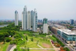 Aerial view of a cityscape featuring modern skyscrapers surrounded by lush green parks. A river curves through the city, lined with both contemporary and traditional buildings. The scene captures the juxtaposition of urban development and natural landscapes.