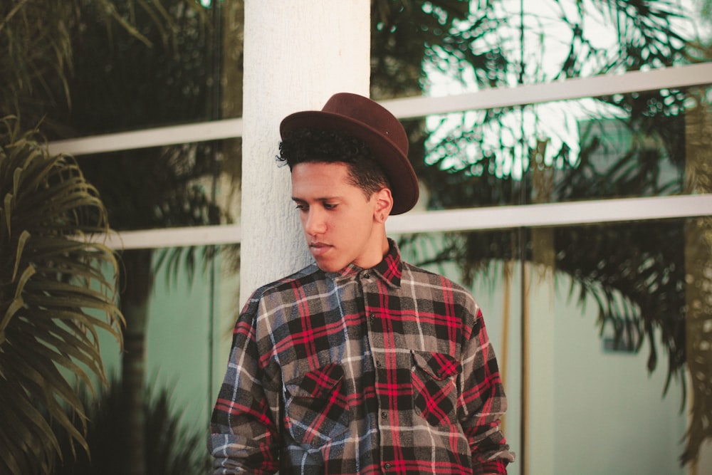 man wearing brown fedora hat and plaid dress shirt while leaning on white post