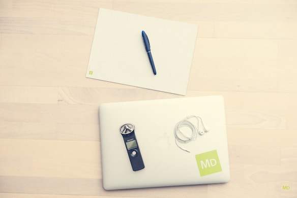 A neatly arranged setup on a light wooden surface, featuring a white laptop with an 'MD' label, white earphones, a black digital recorder, and a blank grid paper with a blue pen placed on top.