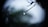 A close-up of a single drop of water hanging from the tip of a thin, bare branch. The background is blurred and appears to be mostly green with some hints of blue and gray, providing a soft and tranquil atmosphere.