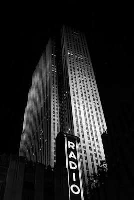 A tall, illuminated skyscraper dominates the night skyline with a vertical sign reading 'RADIO' prominently displayed in the foreground. The structure exudes an Art Deco architectural style, accentuated by the contrast of bright lights against the dark night sky.