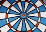 A close-up view of an electronic dartboard with a circular pattern. It features sections of blue, white, and red colors, divided by black plastic lines. The dartboard surface has small perforations for receiving darts.