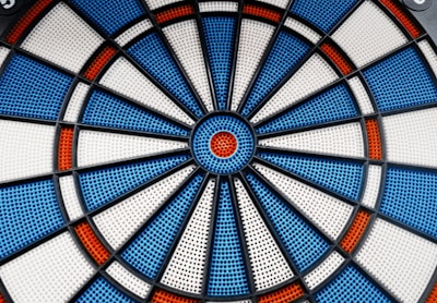 A close-up view of an electronic dartboard with a circular pattern. It features sections of blue, white, and red colors, divided by black plastic lines. The dartboard surface has small perforations for receiving darts.