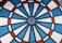 A close-up view of an electronic dartboard with a circular pattern. It features sections of blue, white, and red colors, divided by black plastic lines. The dartboard surface has small perforations for receiving darts.