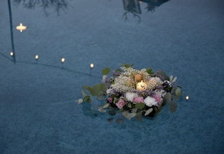 A candle surrounded by a floral arrangement of various flowers and leaves gently floats on the surface of a body of water. Several small candles also float in the background, casting a warm glow. The reflections in the water create a tranquil atmosphere.