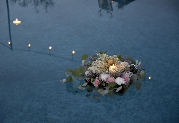 A candle surrounded by a floral arrangement of various flowers and leaves gently floats on the surface of a body of water. Several small candles also float in the background, casting a warm glow. The reflections in the water create a tranquil atmosphere.