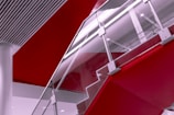 A modern staircase with glass railings and metallic supports, blending into sleek architectural elements. Bold red sections contrast with white and gray tones, creating a dynamic visual composition.