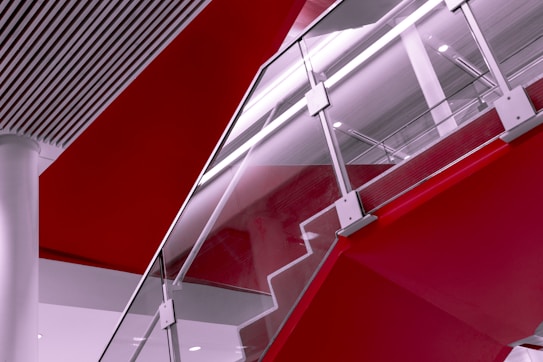 A modern staircase with glass railings and metallic supports, blending into sleek architectural elements. Bold red sections contrast with white and gray tones, creating a dynamic visual composition.